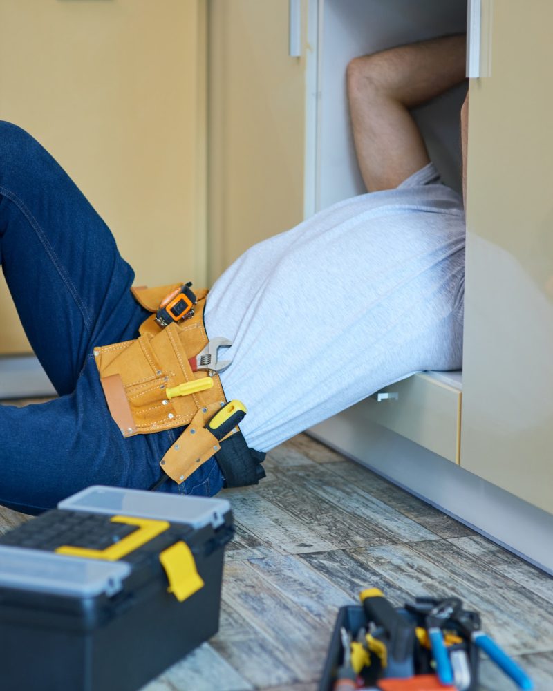 Get fixed. Professional plumber wearing tool belt examining and fixing sink pipe indoors. Tools and Get fixed. Professional plumber wearing tool belt examining and fixing sink pipe indoors. Tools and